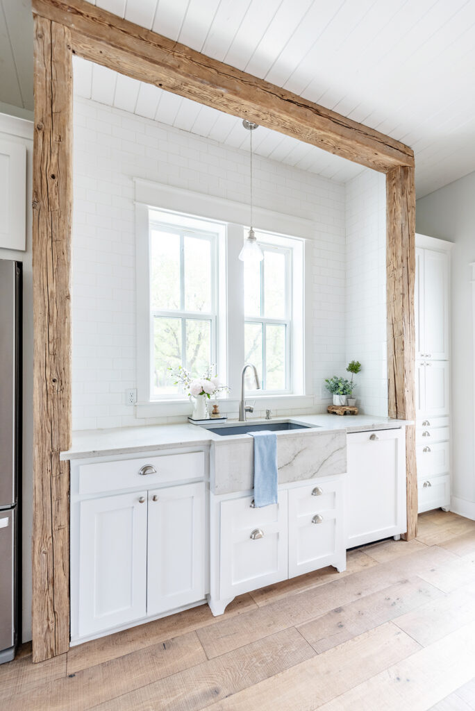 A charming kitchen view of a European farmhouse sink surrounded by white subway tile and reclaimed wood beams and columns. There are 2 windows above the sink and a blue towel draped over the edge.