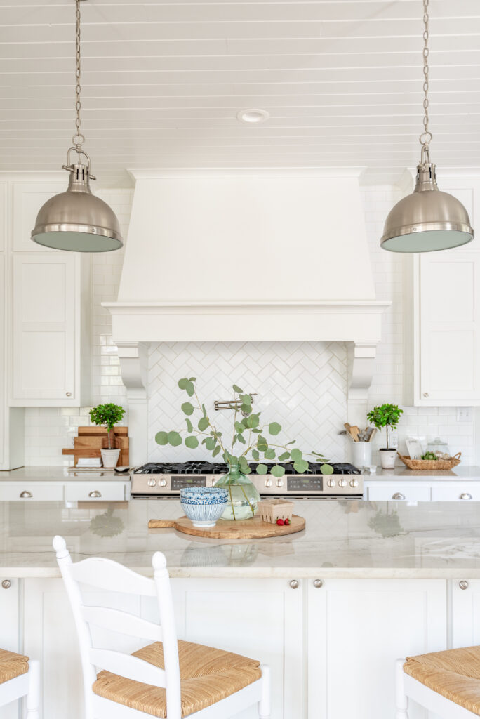 A kitchen with white cabinets and large white vent hood, herringbone subway tile. There's 2 side by side ranges and white quartzite counters