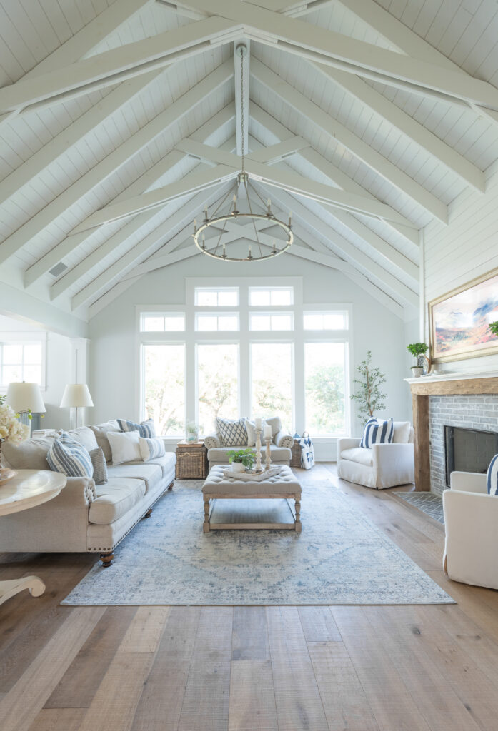 This beautiful living room has vaulted ceilings and exposed rafters with scissor trusses. The ceiling is made of v groove and it is all painted white. The living rooms holds a neutral sofa, upholstered ottoman and an oversized chair. the fireplace has a tv over it that looks like a painting