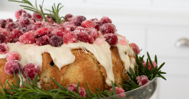 A close up of a bundt cake drizzled with melted white chocolate, sugared cranberries and encircled with fresh sprigs of rosemary.cho