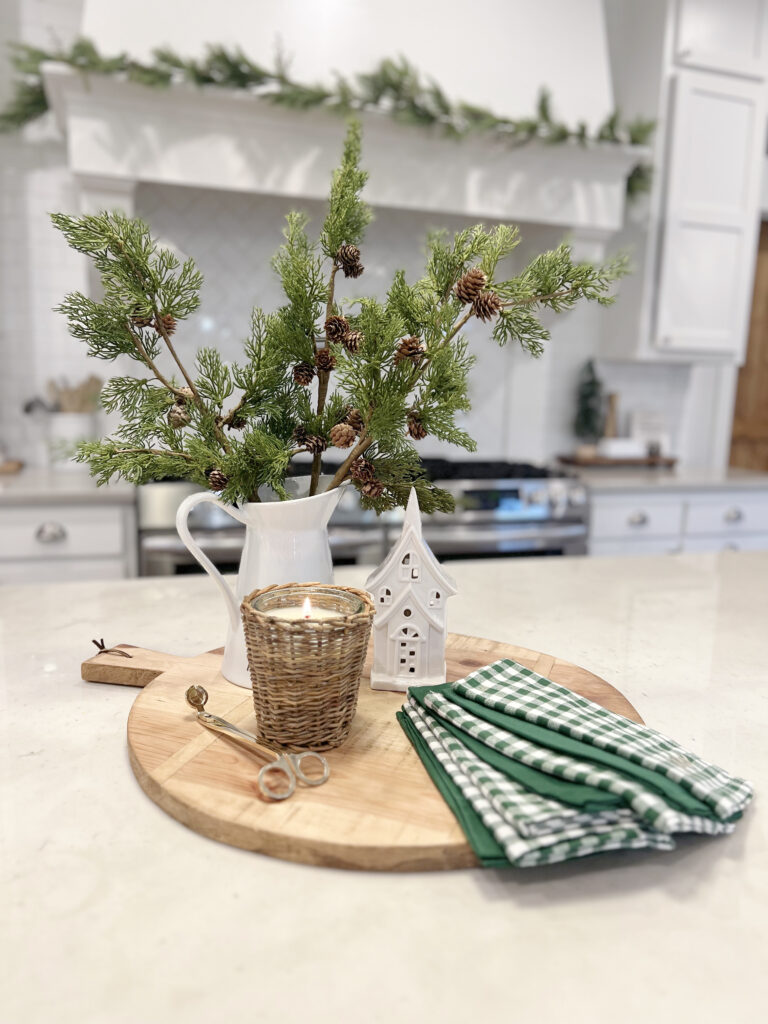 The kitchen island holds a round bread board with a pitcher of pine greenery, a christmas cented candle and some green and white linen napkins.
